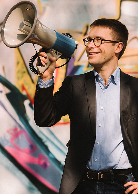 Sebastian Striegel steht vor einer Wand, die mit Graffiti besprüht ist und hält ein Megaphon an seinen Mund.