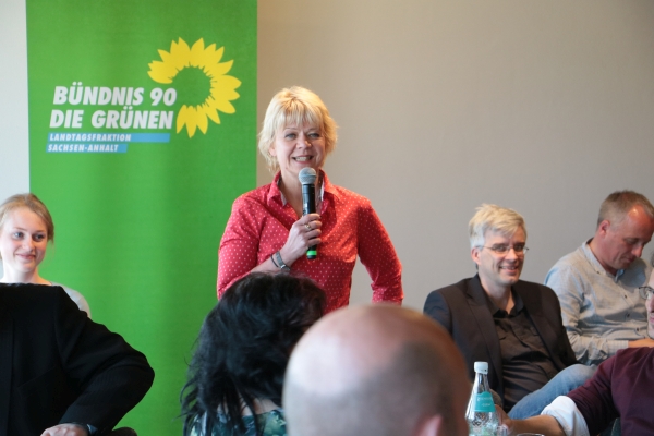 In der Mitte des Bildes steht Cornelia Lüddemann. Sie spricht in einem Mikrophone und lächelt dabei. Sie trägt kurze blonde Haare und eine rote Bluse mit weißen Punkten. Links und rechts von ihr sitzen Personen, die zuhören. 