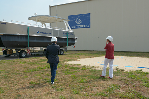 Dorothea Frederking und Sebatian Striegel stehen mit dem Rücken zur Kamera vor einem Boot, der auf einem Tieflader steht. Im Hintergrund ist eine graue Halle zu sehen. Darauf das blaue Logo der Firma Kiebitzberg.