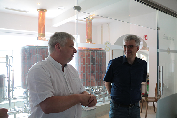 Olaf Meister diskutiert mit einem Mann in einem Raum. Im Hintergrund sieht man ein anderer Raum, der durch Glasscheibe getrennt ist. Darin befindet sich zwei Behälter aus Kupfer mit einem Rohr, der durch die Decke geht. 