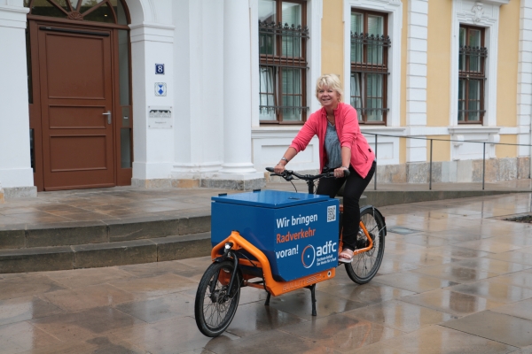 Cornelia Lüddemann ist auf einem orange Lastenrad mit einem blau Kasten. Darauf steht "Wir bringen Radverkehr voran - ADFC". Sie trägt kurze blonde Haare, ein rosanes Fleece-Pullover und eine schwarze Hose und lächelt in die Kamera. Im Hintergrund ist der Eingang vom Landtag zu sehen. 