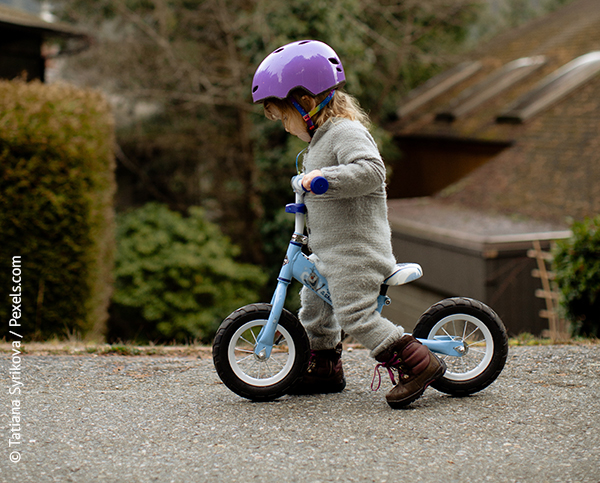 Ein Kind, etwa zwei Jahre alt, steht auf einem blauen Laufrad. Es trägt ein violetter Helm, ein grauer Anzug und braune Schneestiefel. Im Hintergrund sind Häuser und Hecken zu sehen. 