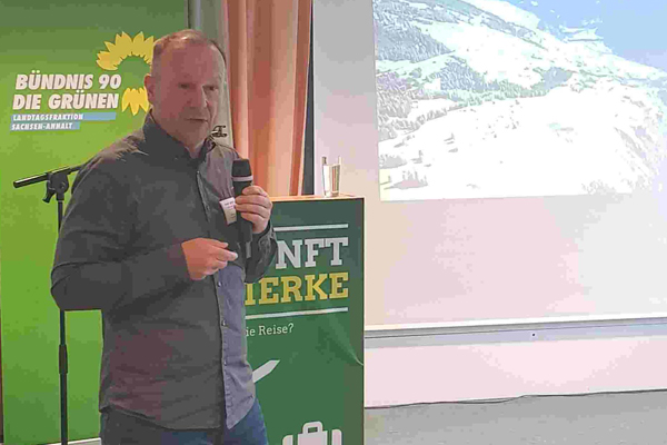 Stefan Forster steht mit dem Mikrofon in der Hand vor dem Redepult und hält seinen Vortrag. Auf der Leinwand ist ein Panorama mit Schnee zu sehen. neben der Leinwand steht ein Aufsteller mit dem Logo der Landtagsfraktion BÜNDNIS 90/DIE GRÜNEN.